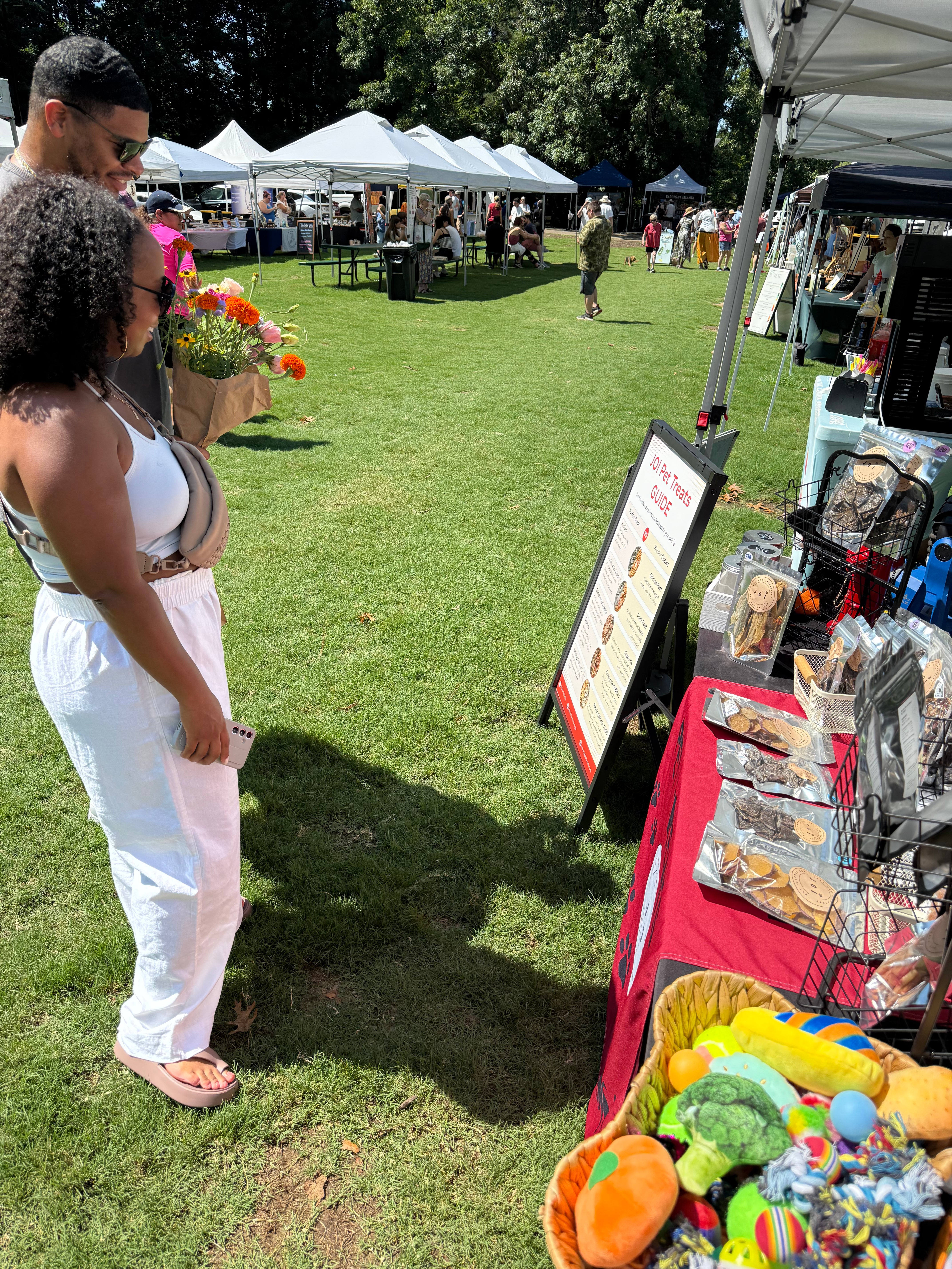 A sunny outdoor market scene on a grassy lawn with white tents, people, and a vendor table in the foreground selling colorful plush toys and other items. Two people are visible on the left, and the market setup fills the frame.