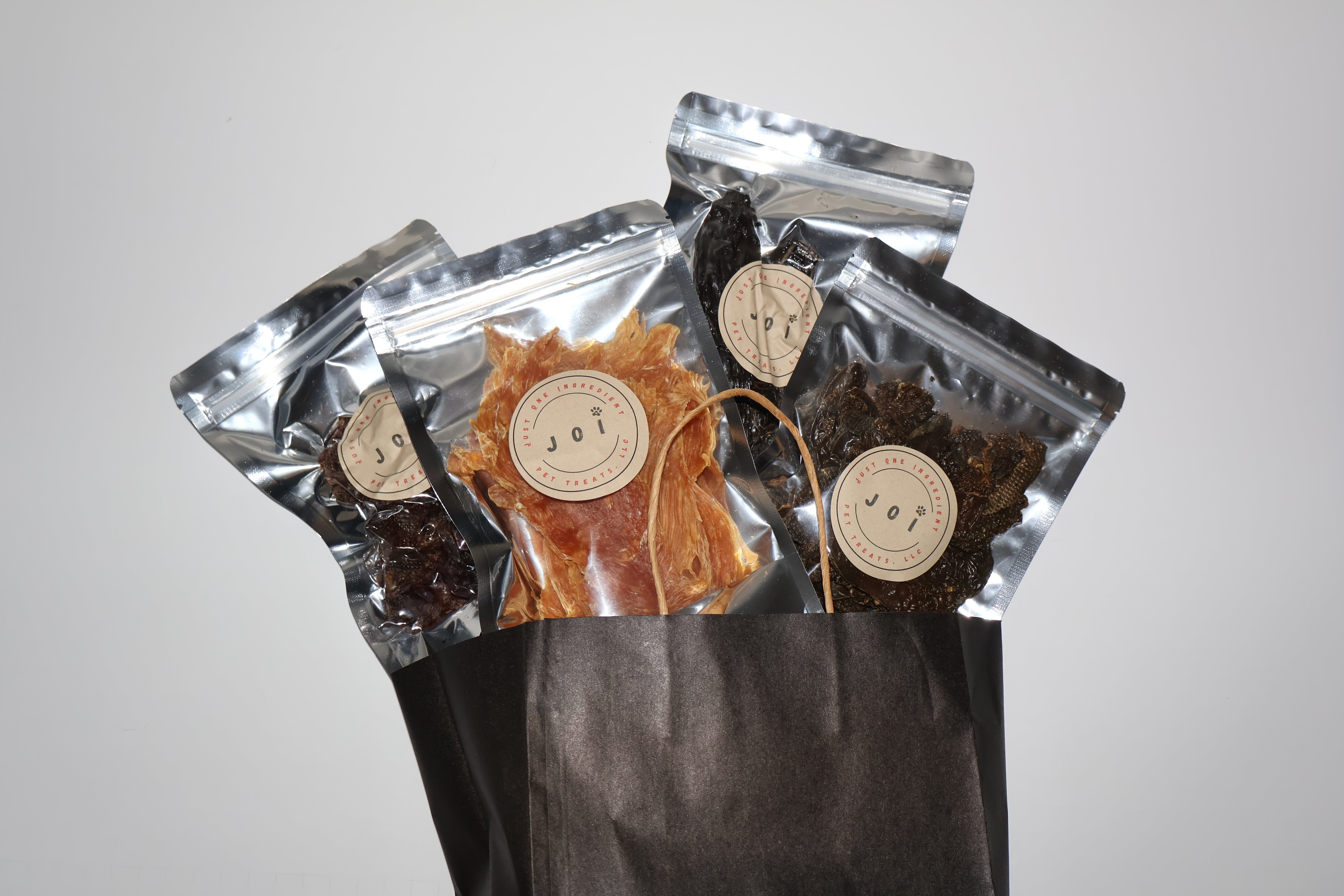 A set of four 3-oz treat bags in a black paper bag, featuring silver resealable pouches with round beige labels and visible treat contents in amber, dark brown, and black tones. Chicken breast, chicken liver, chicken gizzards, and beef liver